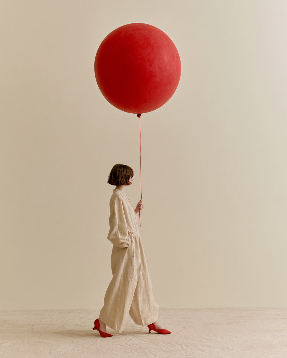 Stillsam modeposter med kvinna i linne som håller en stor röd ballong mot ljus bakgrund.